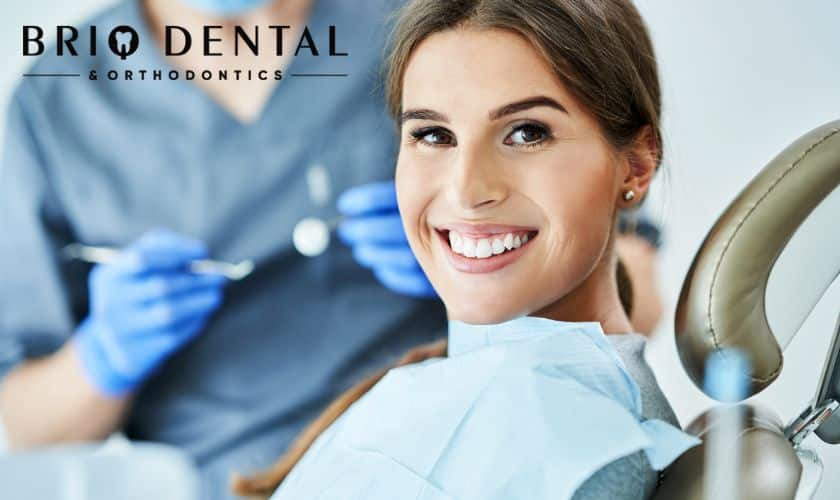 Smiling woman in dental chair during checkup with dentist and equipment visible in background.