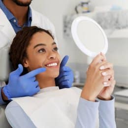 Woman examining her smile in mirror during dental checkup with dentist in background.