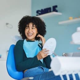 Woman in dental office smiling while speaking on phone, wearing blue scrubs near modern clinic interior.
