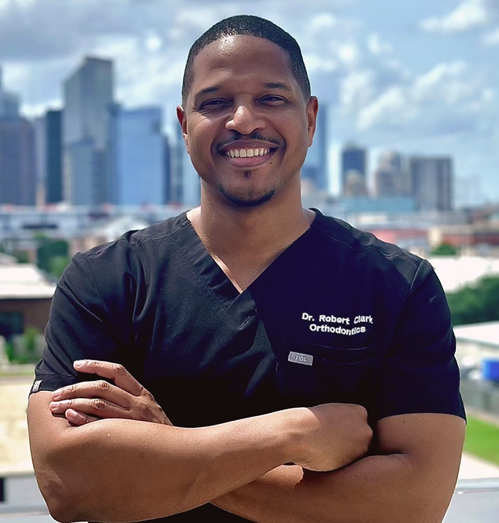 Male dental professional in dark scrubs smiling with arms crossed, city skyline background.