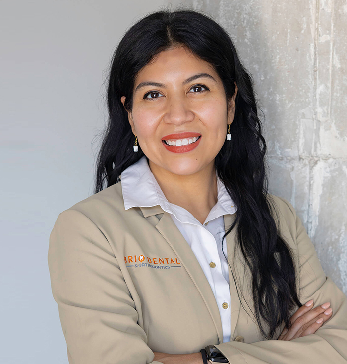 Professional dental staff member in beige uniform smiling with arms crossed against neutral background.