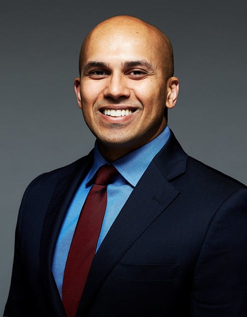 Professional headshot of smiling man in dark blazer and blue shirt against gray background.